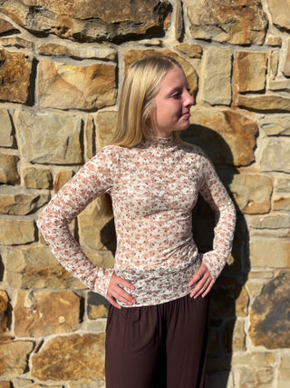 A young blonde woman wearing a long-sleeve mock neck top with a small pink and green floral pattern on a cream background, paired with dark brown pants, standing in front of a rustic stone wall.