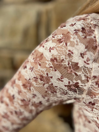 Close-up of a person's arm wearing a sheer floral lace top in cream and dusty rose, with a blurred stone wall background.