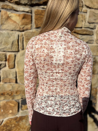 A woman viewed from behind wearing a fitted long-sleeve top with a dense small floral print in muted pink, beige, and brown tones, paired with dark brown bottoms, standing against a stone wall.