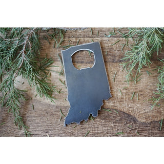 A dark gray metal bottle opener shaped like the state of Indiana, displayed on a rustic wooden surface with green pine branches.