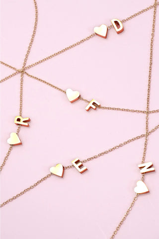 A gold-toned chain necklace with alternating heart charms and letter charms spelling out a name, laid out on a solid light pink background.