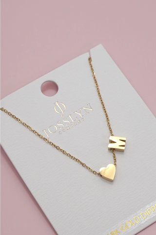 A gold necklace with a letter 'M' pendant and a heart charm, displayed on a white Josslyn Gold Cards brand card against a soft pink background.