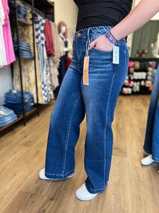 A person wearing a pair of wide-leg blue denim jeans in a clothing store, standing on a wood floor with racks of clothes in the background.