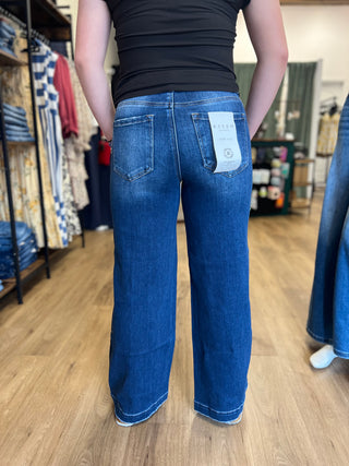 A person standing in a clothing store, viewed from behind, wearing blue wide-leg jeans with a white paper tag attached to the back pocket.