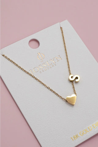 A gold necklace with a heart pendant and a letter 'S' initial pendant, displayed on a white jewelry card with the brand name JOSSLYN CO., which is placed on a soft pink background.