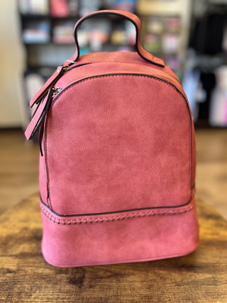 A pink faux-leather mini backpack with a top handle, shown on a wooden table with a blurred store background.