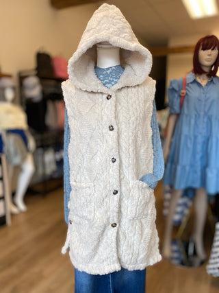A cream-colored, textured hooded vest with front buttons, displayed on a mannequin torso in a clothing store. The vest is layered over a light blue patterned long-sleeve top and blue jeans.
