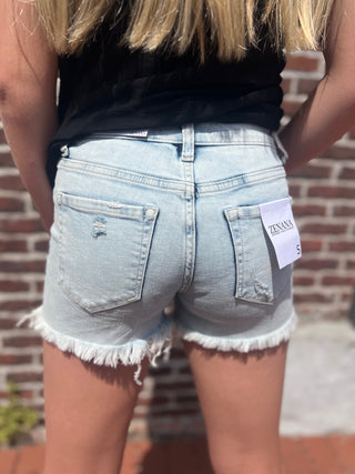 Person wearing light blue denim shorts with a brand tag, standing against a brick wall.