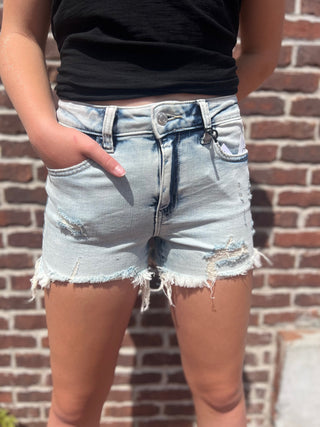 Person wearing light blue denim shorts with a black top against a brick wall.
