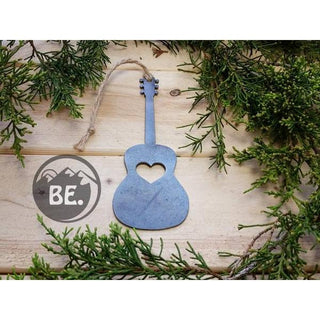 A rustic  guitar-shaped ornament with a heart-shaped cutout in the center, hanging from a jute twine loop. The ornament is displayed on a light wood background surrounded by green pine branches, with a circular 'BE.