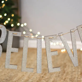 Silver glittery 'BELIEVE' banner with blurred Christmas lights in the background