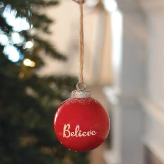 Red Christmas ornament with 'Believe' text hanging against a blurred festive background