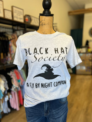 A light gray heather graphic t-shirt displayed on a dress form mannequin inside a retail store. The shirt features a black printed graphic that reads 'BLACK HAT Society' and 'A FLY BY NIGHT COMPANY' with a witch hat silhouette.