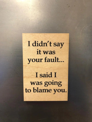 A rectangular light wood sign with black printed text that reads, 'I didn't say it was your fault.