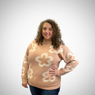 A woman with curly brown hair wearing a blush knit sweater with large cream-colored floral motifs, paired with dark blue jeans, standing against a light gray studio background.