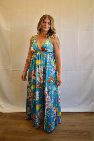 A woman modeling a blue floral maxi dress with a tie waist, standing against a white fabric backdrop.