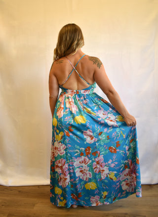 A woman viewed from behind wearing a blue maxi dress with a colorful floral print and thin crisscross back straps, standing against a cream fabric backdrop.
