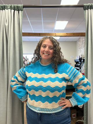 A smiling woman with curly brown hair stands in a fitting room wearing a hand-knit crewneck sweater with wavy horizontal stripes in turquoise blue and cream, paired with blue jeans.