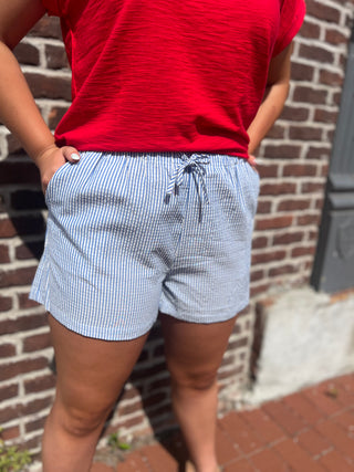 Person wearing a red shirt and blue striped shorts standing against a brick wall.
