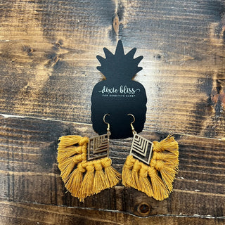 A pair of yellow tassel earrings with geometric gold-tone metal charms, displayed on a black pineapple-shaped card that reads 'dixie bliss gold sensitive ears' against a rustic dark wood background.
