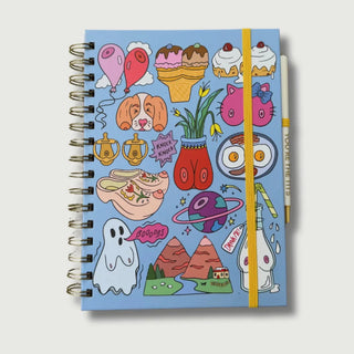 A spiral-bound notebook with a light blue cover covered in colorful whimsical doodles, including ice cream cones, cupcakes, a dog face, a red female silhouette, a ghost, mountains, and a bottle. A yellow elastic closure band wraps around the notebook, and a white pen with a blue cap rests on the right side.