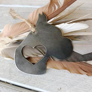 A rustic metal cat-shaped ornament with a heart-shaped cutout, hanging from a twine string and styled with tan and white feathers on a whitewashed wooden surface.