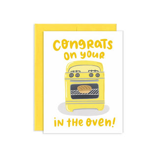 A greeting card with a yellow envelope, featuring a white card front with the text 'Congrats on your oven in the oven!' and a yellow illustration of an oven with a loaf of bread inside.