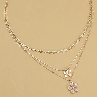 A layered gold-tone necklace featuring a delicate chain, a small butterfly pendant, and a larger four-leaf clover pendant embellished with clear rhinestones, displayed on a solid beige background.