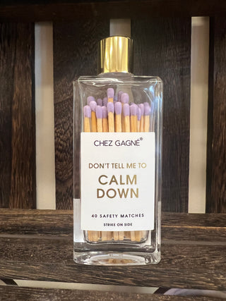 A clear rectangular glass bottle of matchbox incense sticks with a gold cap and a white label reading 'CHEZ GAGNÉ' and 'DON'T TELL ME TO CALM DOWN', placed on a dark wooden slatted surface.