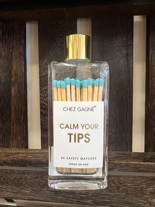A clear rectangular glass bottle filled with safety matches, featuring a white label that reads 'CHEZ GAGNÉ CALM YOUR TIPS' and a shiny gold cap, displayed on a dark wooden slatted surface.