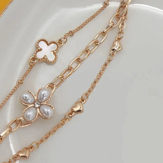 A close-up of a layered gold-tone bracelet set on a white plate. The necklace features a delicate cable chain, a fine cable chain, and a charm link chain, all adorned with white faux-pearl flower charms and small gold heart charms.