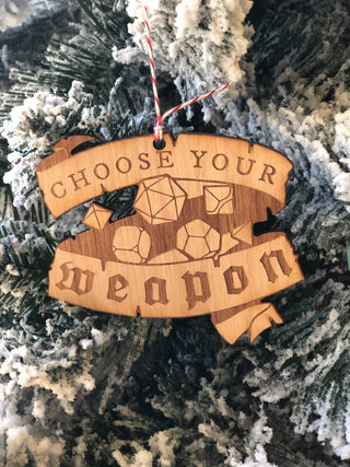 A wooden ornament shaped like a torn scroll with engraved dice and the words 'CHOOSE YOUR WEAPON,' hanging against a snowy tree branch background.