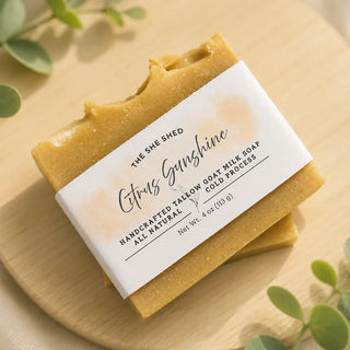 A bar of handmade goat milk soap labeled 'Citrus Sunshine' by The She Shed, resting on a light wooden board with green leaves in the background.