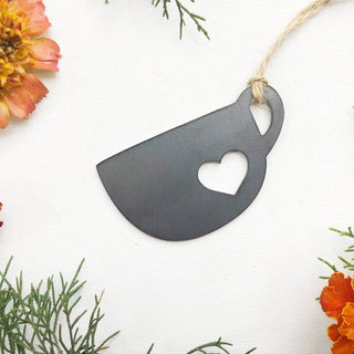 A matte black metal ornament shaped like a teacup with a heart-shaped cutout in the center, hanging from a natural jute twine. The ornament is displayed on a textured white fabric background, framed by decorative orange flowers and green pine branches.