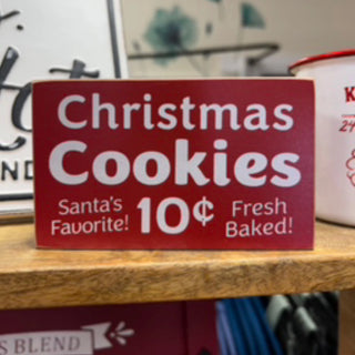 A rectangular red sign on a wooden shelf with white text reading 'Christmas Cookies' and smaller phrases 'Santa's Favorite!' and '10¢ Fresh Baked!'.