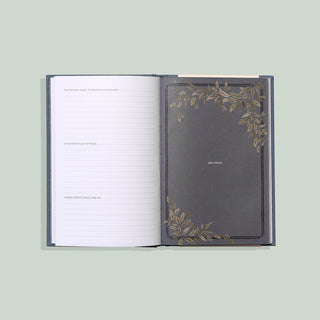 An open hardcover notebook or journal on a pale green background. The left page has lined writing space with a caption above, and the right page shows a dark gray cover decorated with gold-toned botanical corner illustrations and the text 'THE PLANET'.