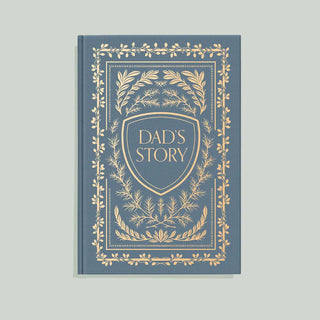 A hardcover book or journal titled 'DAD'S STORY' with a dusty slate blue cover and ornate gold foil detailing, centered on a light gray background.