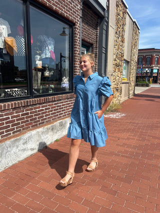Person in a blue dress standing on a sidewalk next to a brick building.