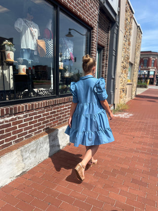 Person in a blue dress walking on a brick sidewalk in front of a store.