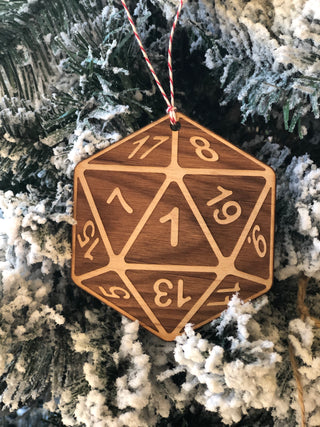 A wooden D20 polyhedral ornament with engraved numbers, hanging from a red and white string against a snowy, frosted Christmas tree.