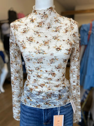 A long-sleeve mock neck top with a dense floral print in beige, brown, and muted orange on a white base, displayed on a mannequin torso with blue jeans. A brand tag is visible at the waist.