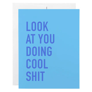 A light blue greeting card with bold dark blue text reading 'LOOK AT YOU DOING COOL SHIT' on a white envelope, shown against a plain white background.