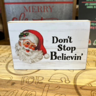A rectangular white wooden sign with a vintage-style Santa face on the left and the phrase 'Don't Stop Believin'' in black text on the right, displayed on a wooden surface.