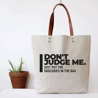 A beige canvas tote bag with brown leather handles, featuring the black printed text 'DON'T JUDGE ME. JUST PUT THE GROCERIES IN THE BAG'.