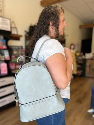 A woman in a white ribbed short-sleeve top and blue jeans is shown from the side/back in a shop setting, wearing a light sage green faux-leather backpack with a top handle and zipper closure.