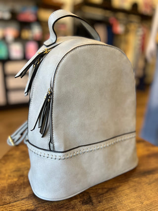 A light gray faux-leather backpack with black leather tassel zippers and a top handle, resting on a wooden table in a store setting.