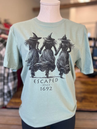 A light mint green short-sleeve t-shirt displayed on a mannequin torso in a store setting. The shirt features a black graphic of three witches holding hands, with the text 'ESCAPED SINCE 1692' printed below.