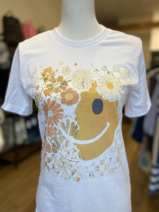 A white short-sleeve graphic T-shirt displayed on a mannequin torso in a retail setting. The front graphic features a large mustard-yellow smiley face surrounded by orange, beige, and white flowers, daisies, checkerboard patterns, and paint splatter details.