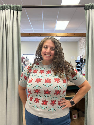 A woman with curly brown hair stands in a fitting room wearing a light gray short-sleeve knit top with a red and green floral pattern, paired with blue jeans.