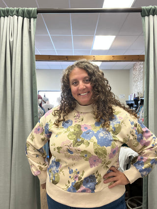 A woman with curly brown hair stands in a fitting room wearing a cream-colored sweatshirt with a colorful floral pattern in blue, pink, and green, paired with blue jeans.